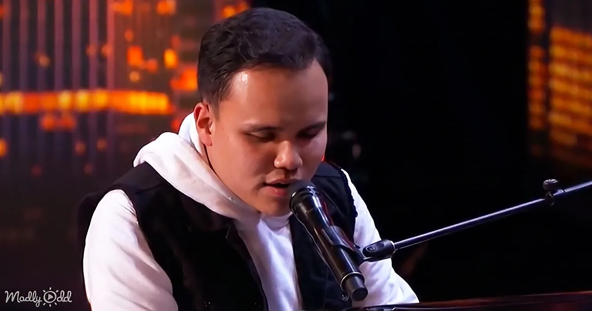 A man in a white hoodie and black vest sings into a microphone on stage with a blurred background.
