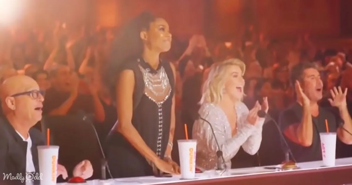 A woman in a black dress stands at a table with judges, clapping and cheering in front of a crowd.