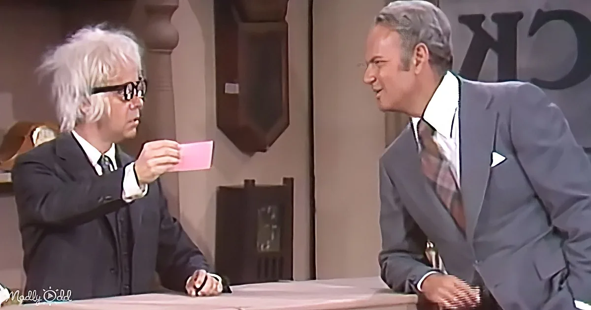 A man in a suit holds a red card while another man in a suit leans forward, both seated at a table.