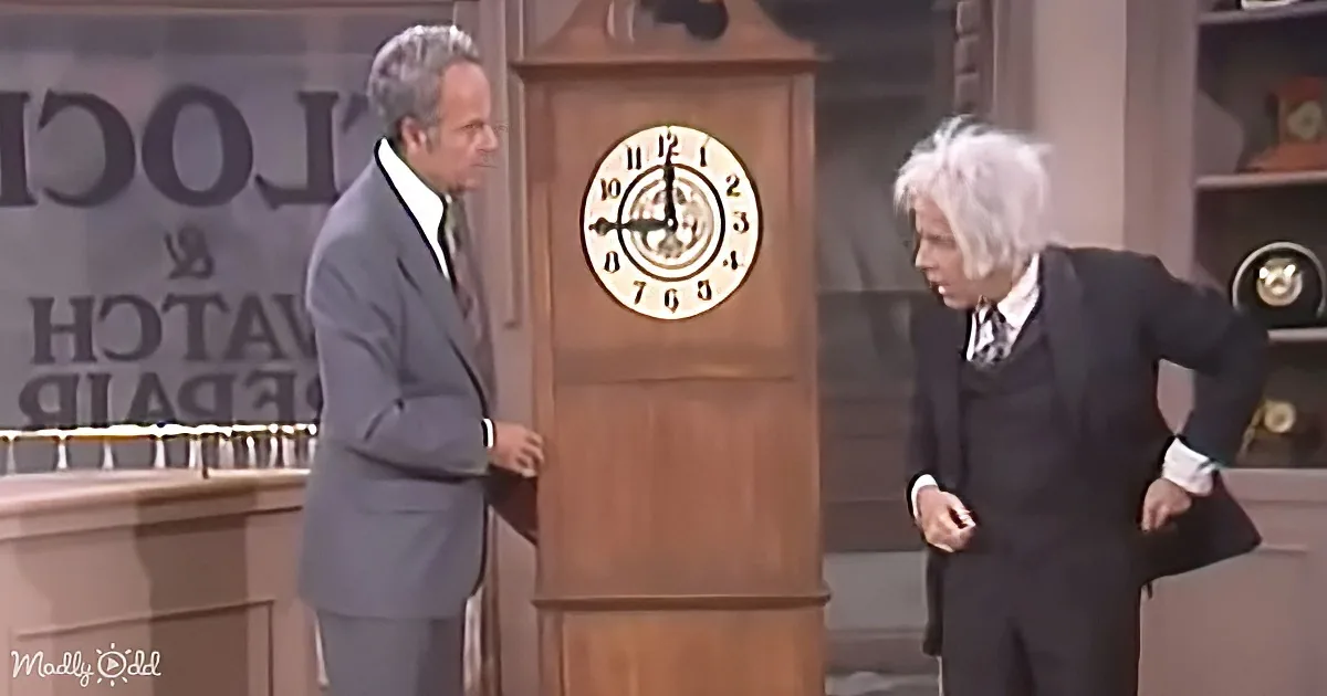 Two men in suits stand beside a large grandfather clock on a stage set.