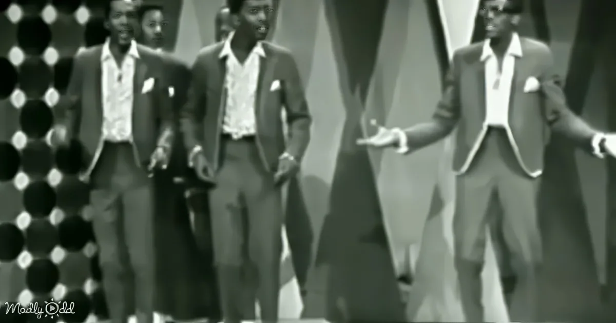 Four men in suits and white shirts perform on stage with a geometric background.