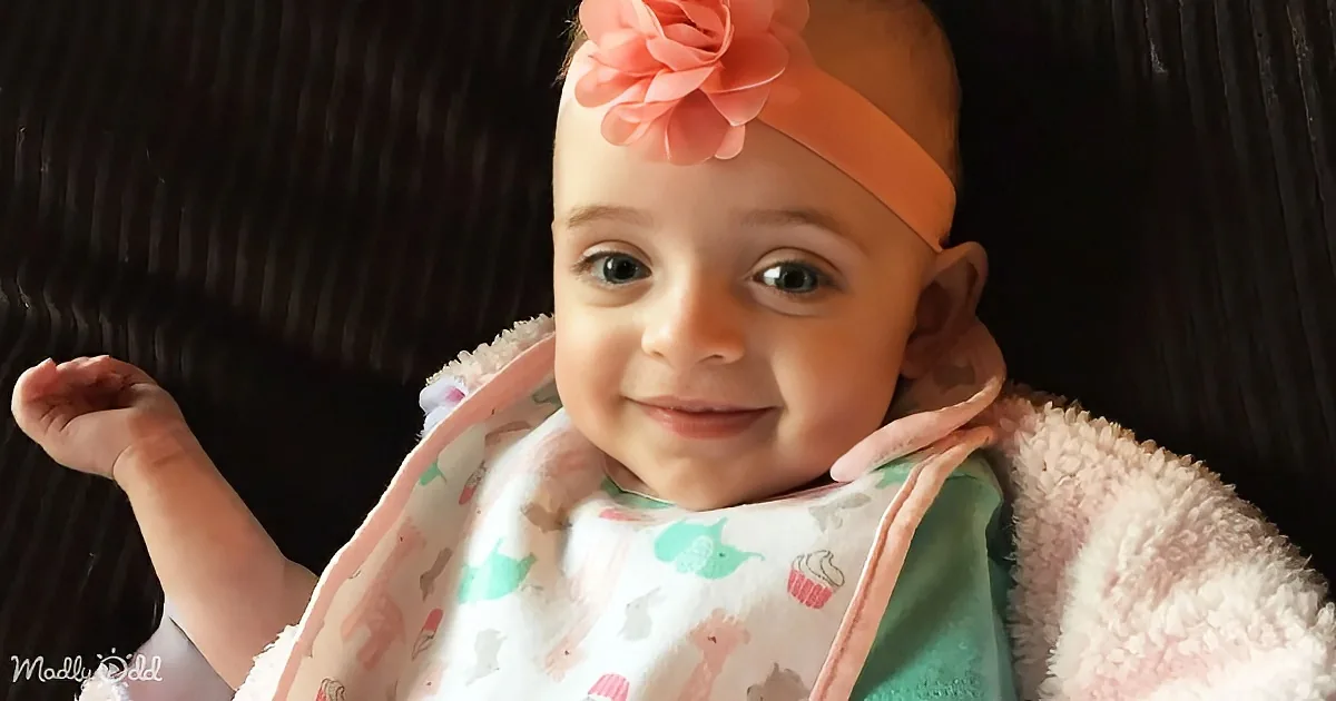 A smiling baby wearing a pink flower headband and a bib with colorful designs, sitting on a dark background.