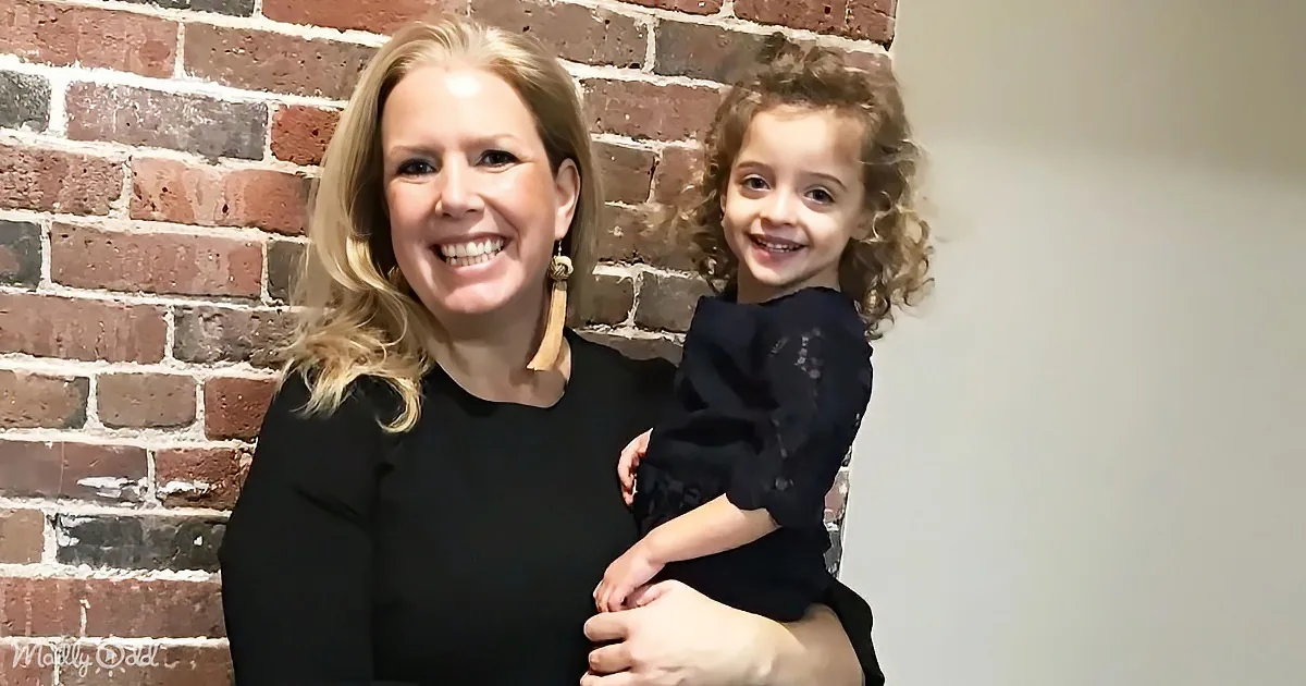 A woman holds a smiling child with curly hair against a brick wall backdrop.