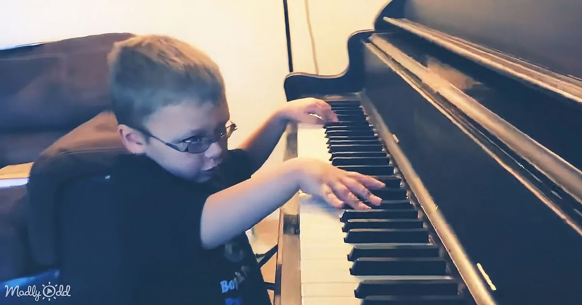 A boy wearing glasses plays a piano, his hands positioned on the keys.