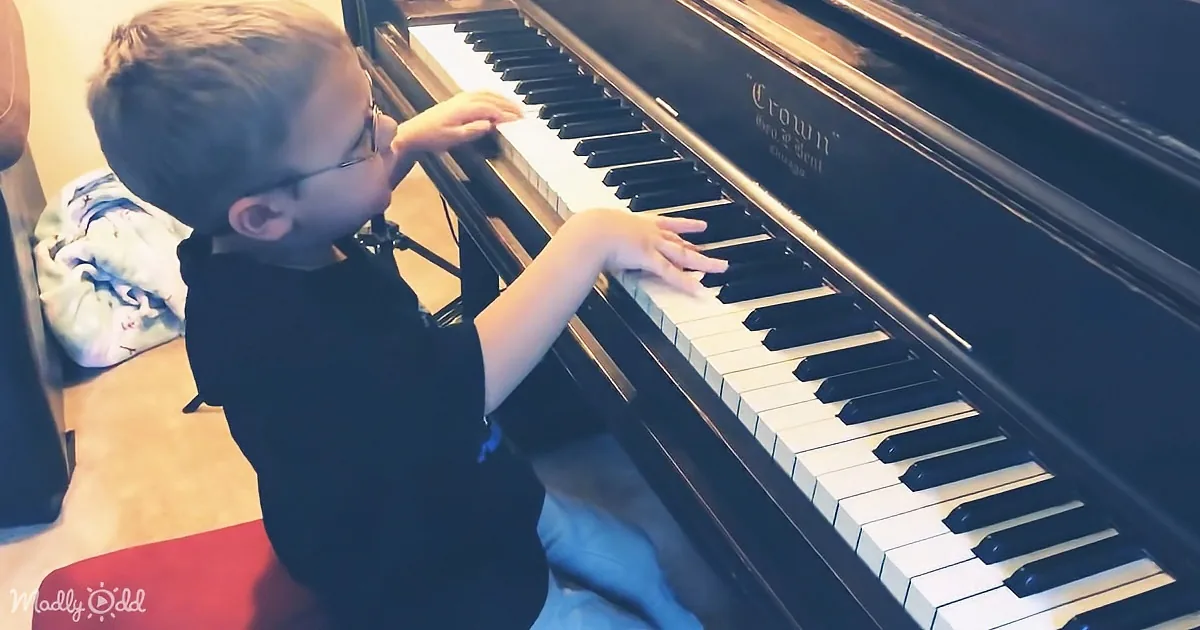 A young boy wearing glasses plays a black Crown piano, focusing intently on the keys.
