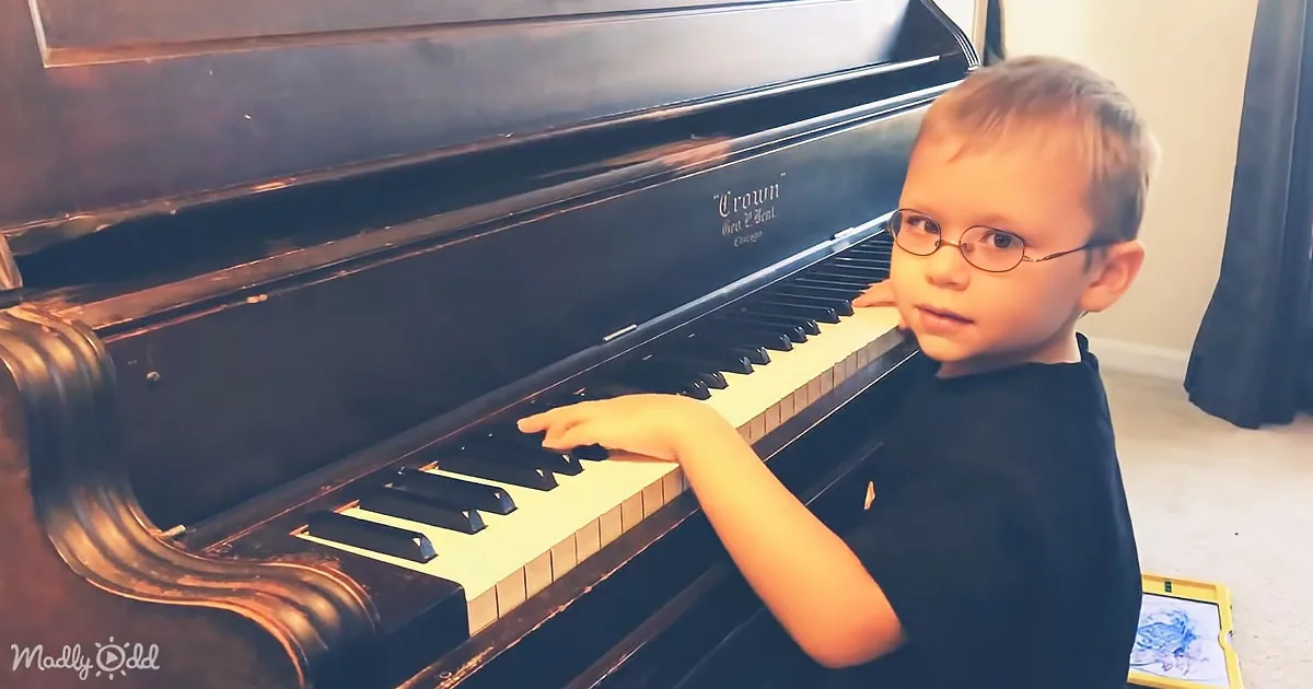 A young boy wearing glasses plays a black piano, focusing on the keys.