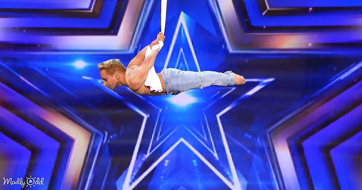 A man performs an aerial trick, hanging from a rope with a star-shaped background.
