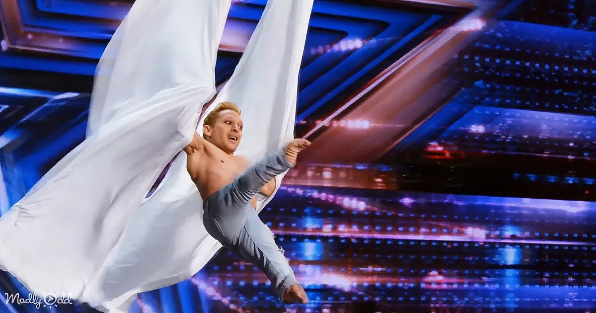 A shirtless man performs aerial acrobatics with flowing white fabric against a vibrant stage backdrop.