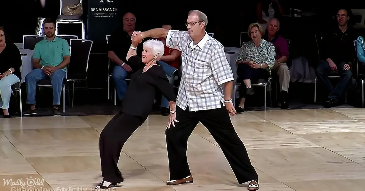An elderly couple dances gracefully on a wooden floor, surrounded by spectators in a dimly lit venue.