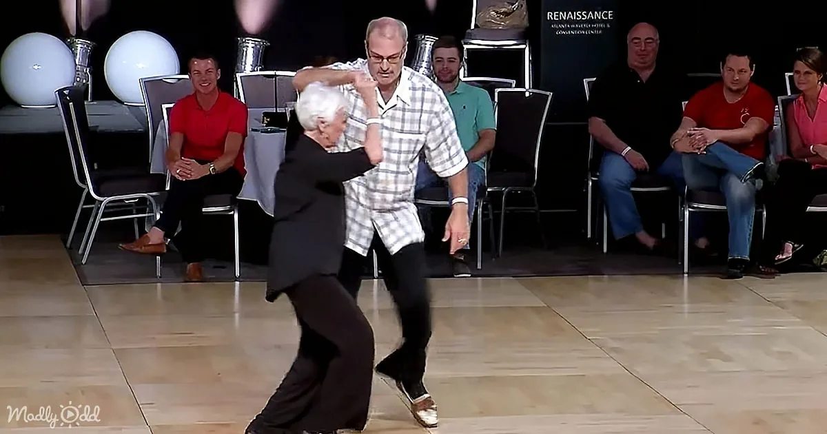 An elderly couple dances gracefully on a wooden floor, surrounded by seated spectators.