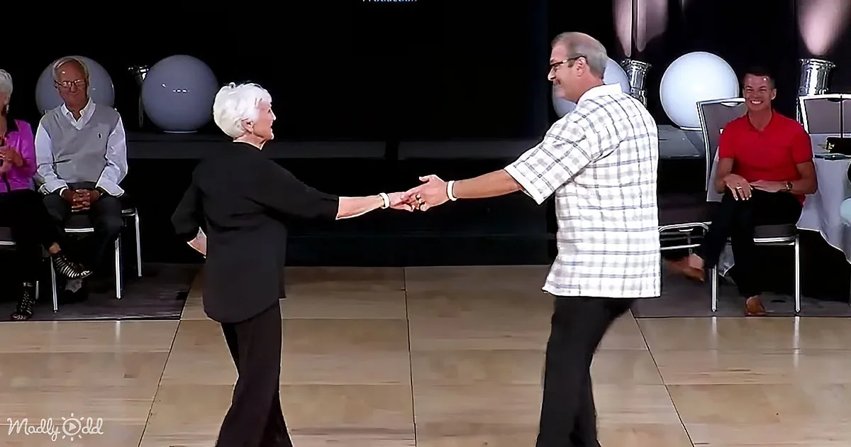 An older couple dances hand-in-hand on a stage, surrounded by seated spectators.