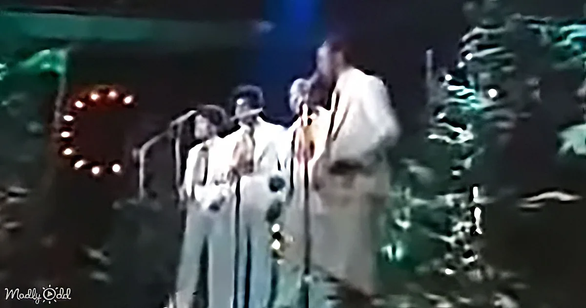 A group of men in suits and hats sing on stage with microphones, surrounded by Christmas decorations.