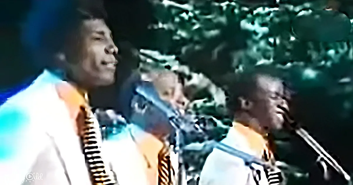 A man in a white suit sings into a microphone on stage, surrounded by other musicians.