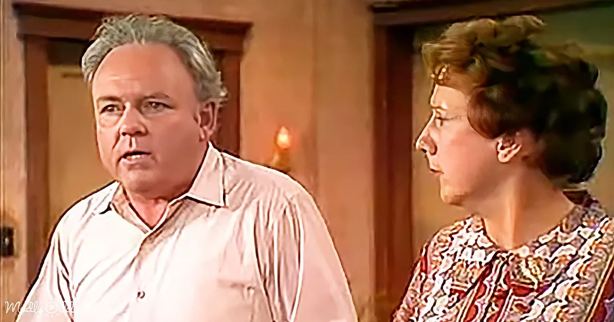 A man in a white shirt stands next to a woman in a patterned blouse, both facing each other in a room with wooden doors.