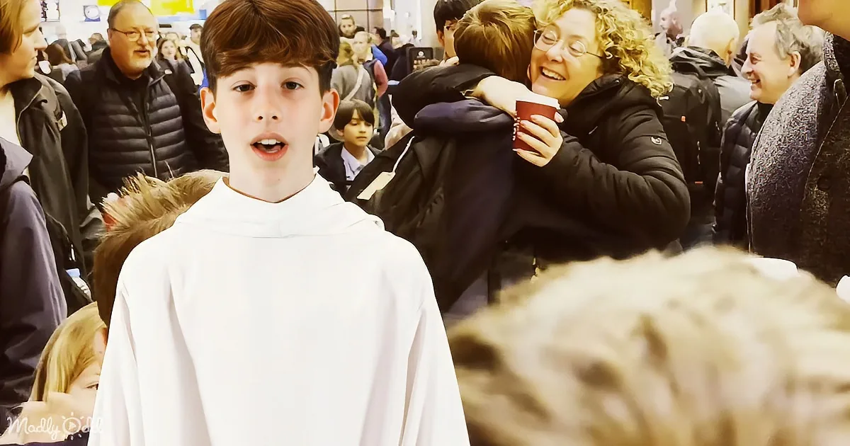 A boy in a white robe stands in a crowded area, smiling, with people hugging and holding cups in the background.
