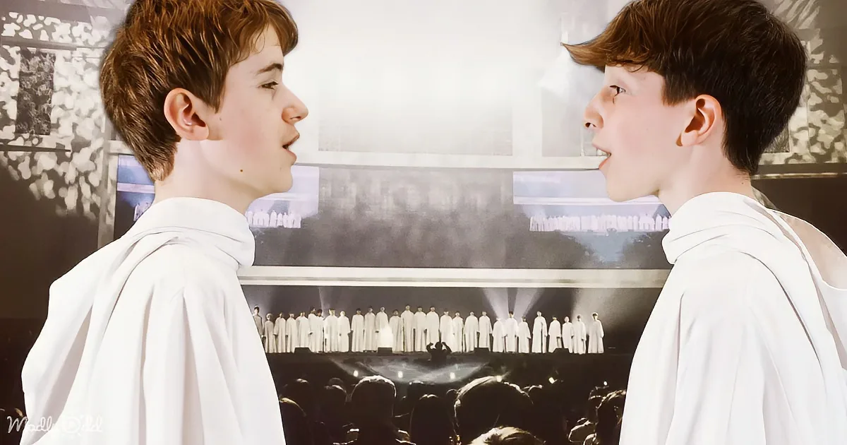 Two young boys in white robes face each other, with a choir and audience in the background.