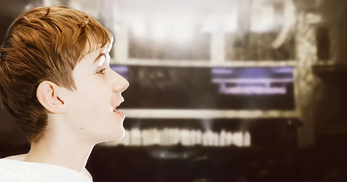 A young person with short brown hair is singing, mouth open, in a dimly lit room with shelves in the background.