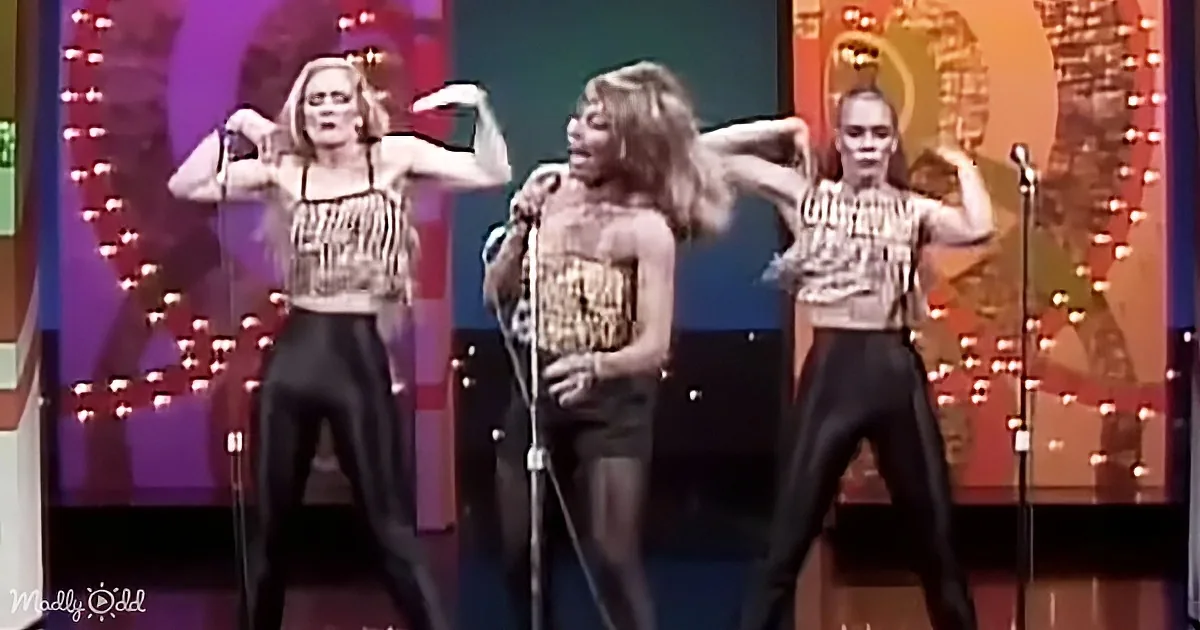 A woman in a gold dress sings into a microphone, flanked by two dancers in black pants and patterned tops.