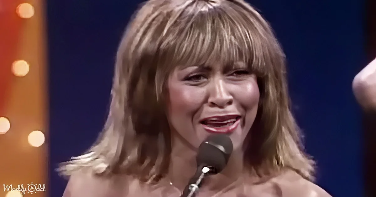 A woman with shoulder-length blonde hair and bangs is singing into a microphone.