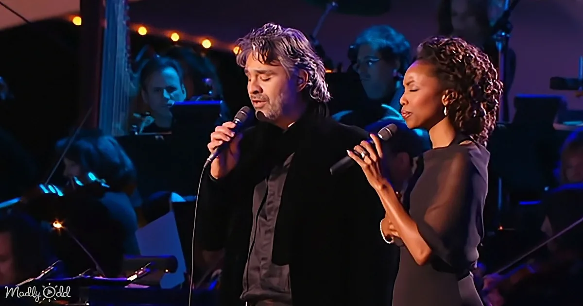 A man and woman sing into microphones on stage, surrounded by musicians.