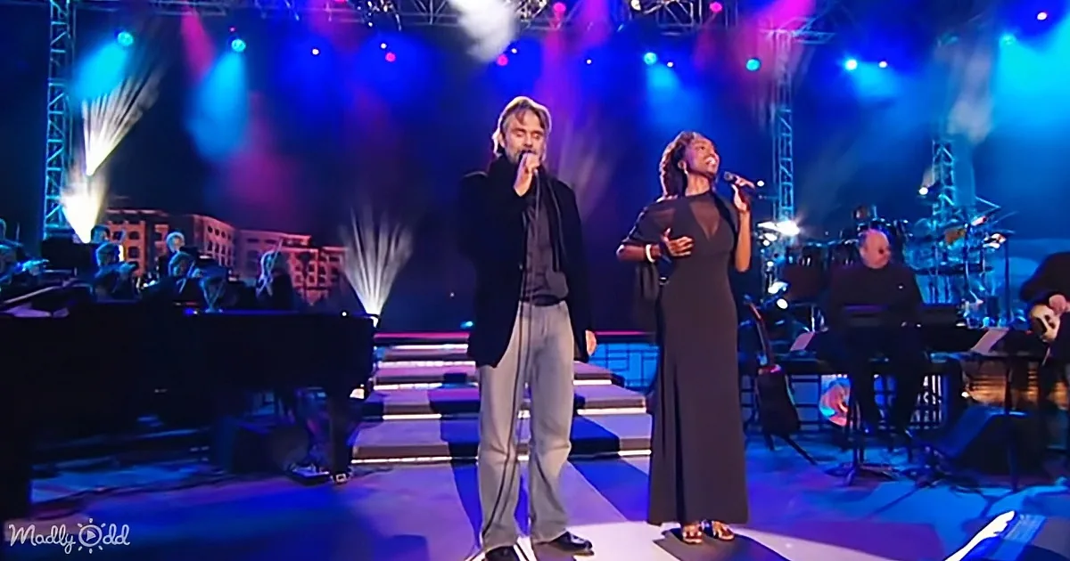 A man and woman sing on stage with a band, vibrant lights and smoke in the background.