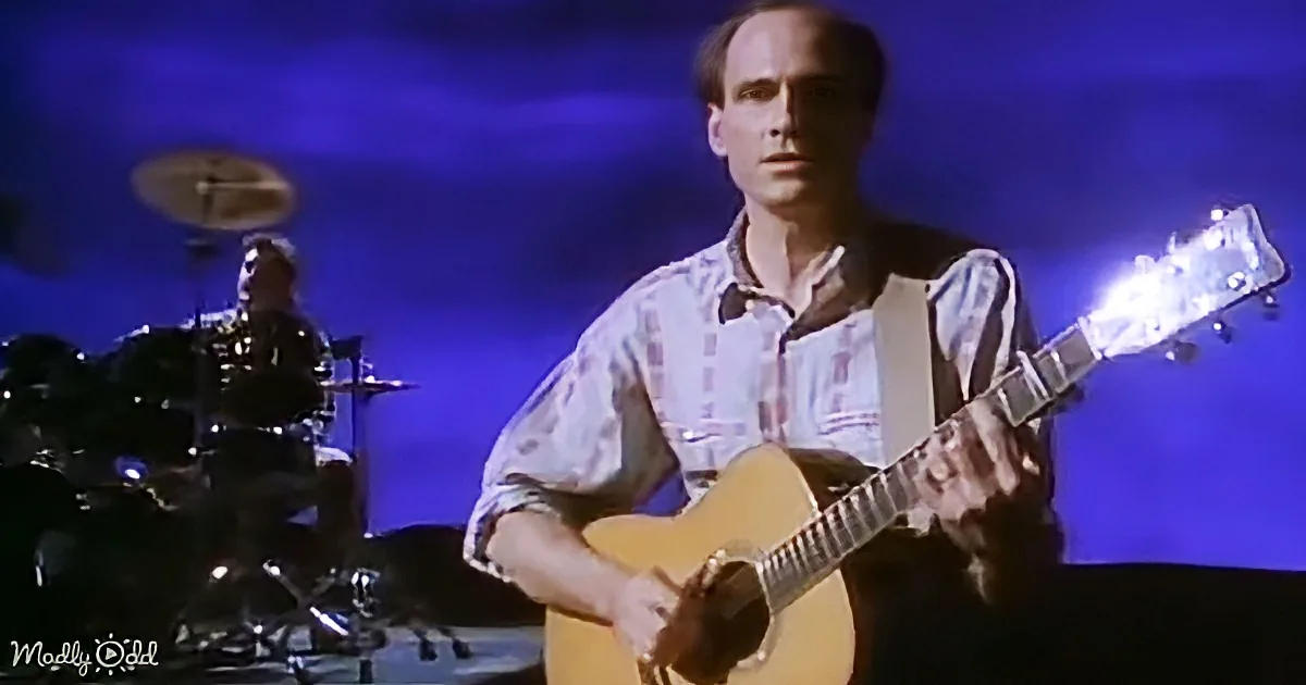 A man plays an acoustic guitar on stage, with a drummer in the background under blue lighting.