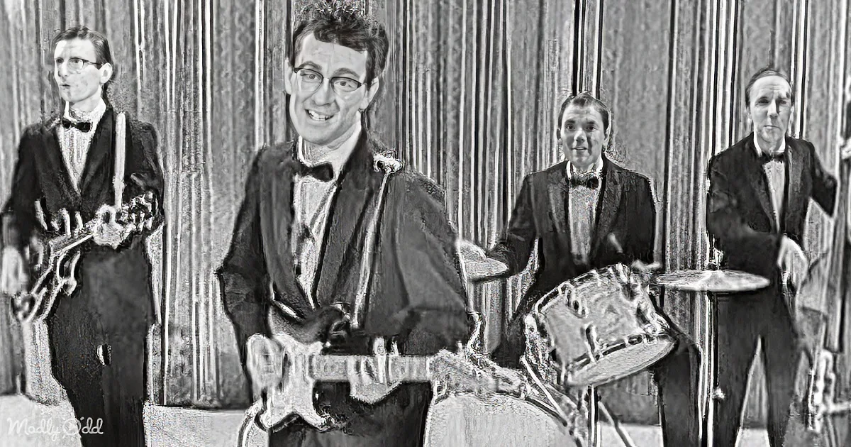 A man in a suit plays guitar, smiling, with a band behind him, including a drummer and a bassist.