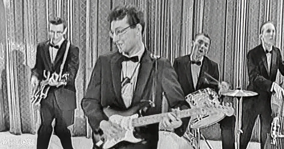 A man in a suit plays guitar while a saxophonist, drummer, and another musician in a tuxedo look on.