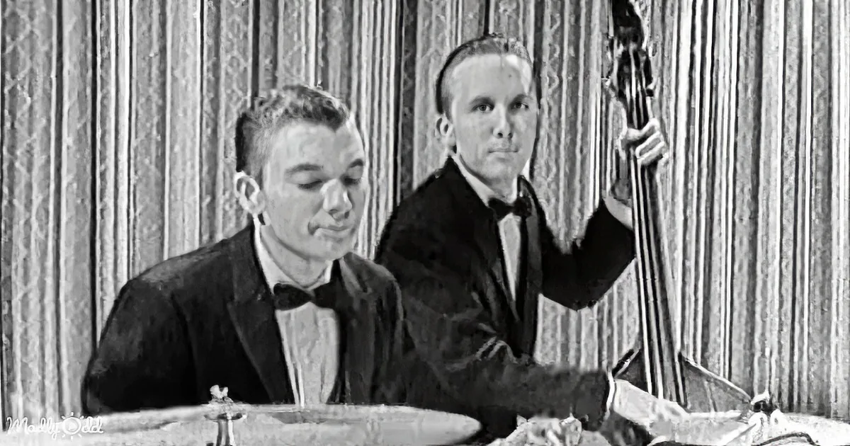 A man in a tuxedo sits at a table, smiling, while another man plays a double bass in the background.