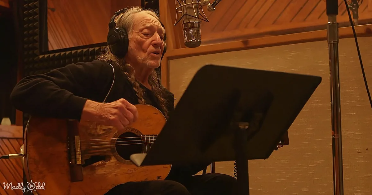 An older man with headphones plays guitar in a recording studio, focused on sheet music.