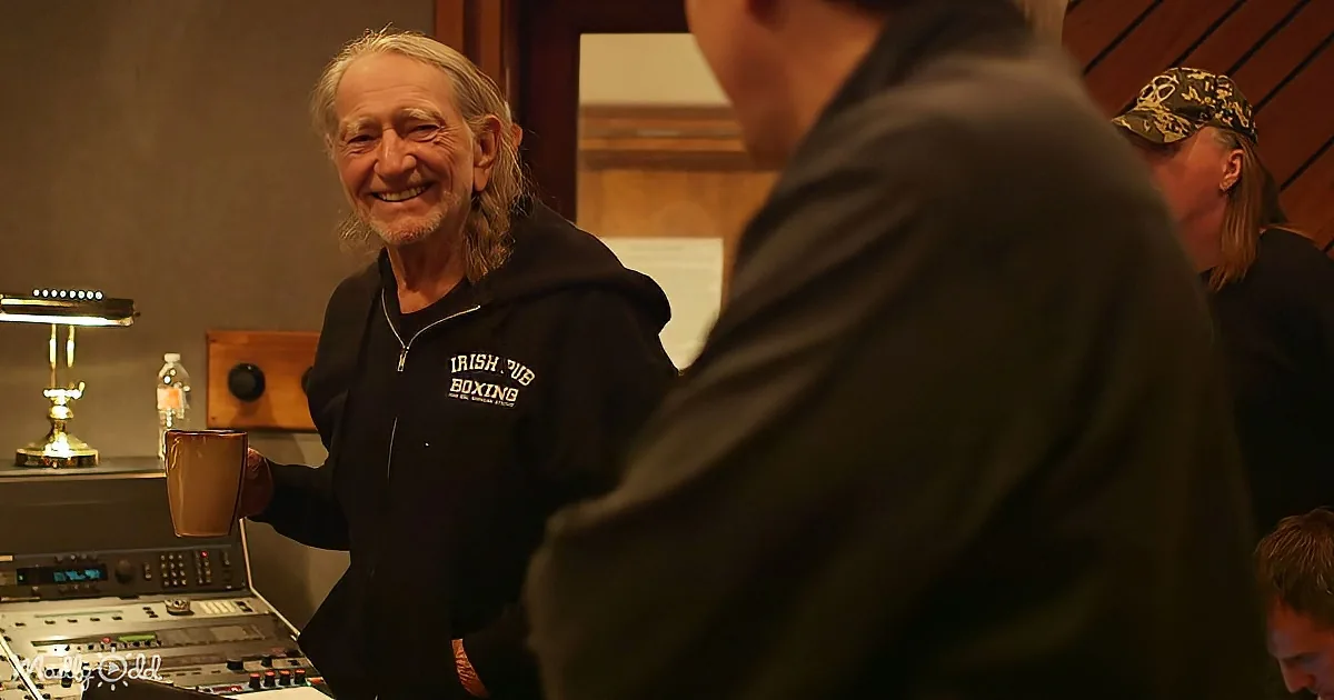 A smiling man in a black hoodie holds a mug, standing in a recording studio with others around.