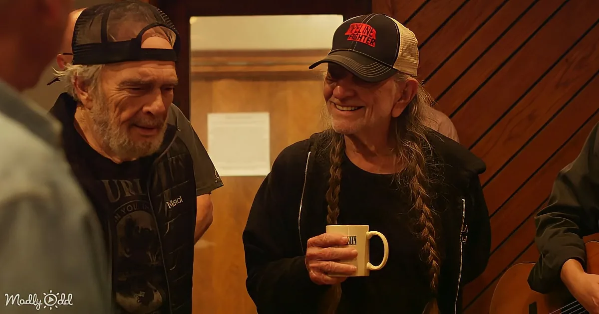 Two men with long hair and hats are smiling and holding a mug, standing in a room with wooden walls.