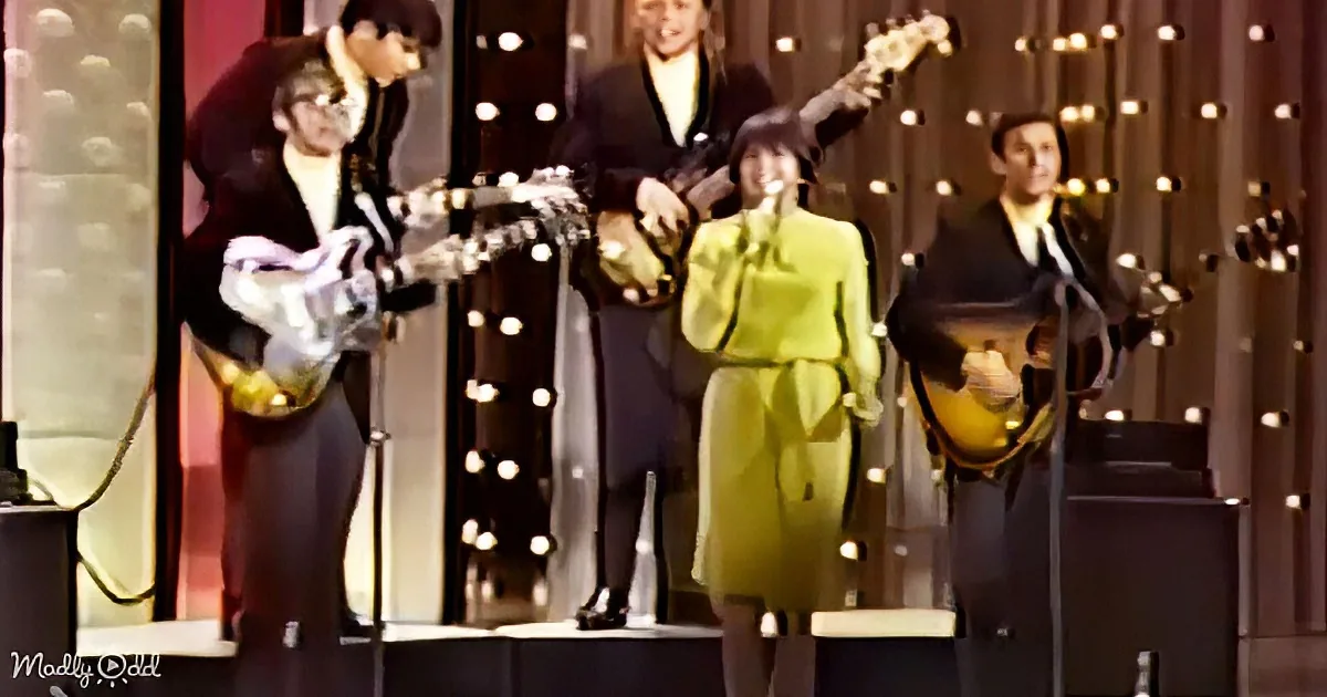 A group of musicians in 1960s attire performs on stage with electric guitars and a microphone.