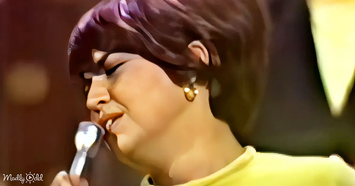 A person with purple hair and gold earrings sings into a microphone, wearing a yellow top.