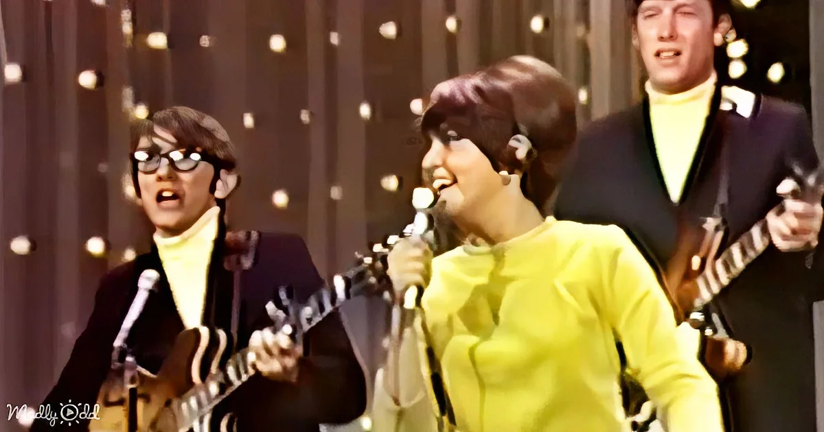 A woman in a yellow outfit sings into a microphone, accompanied by musicians playing guitars.