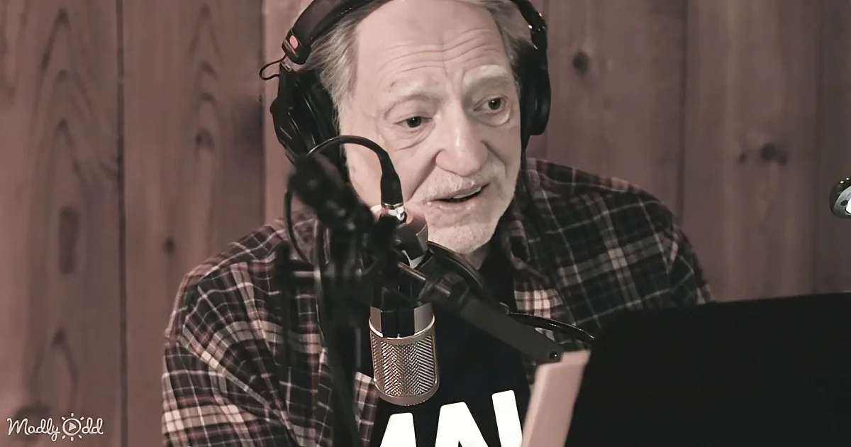 An older man in a plaid shirt speaks into a microphone, wearing headphones, in a studio setting.