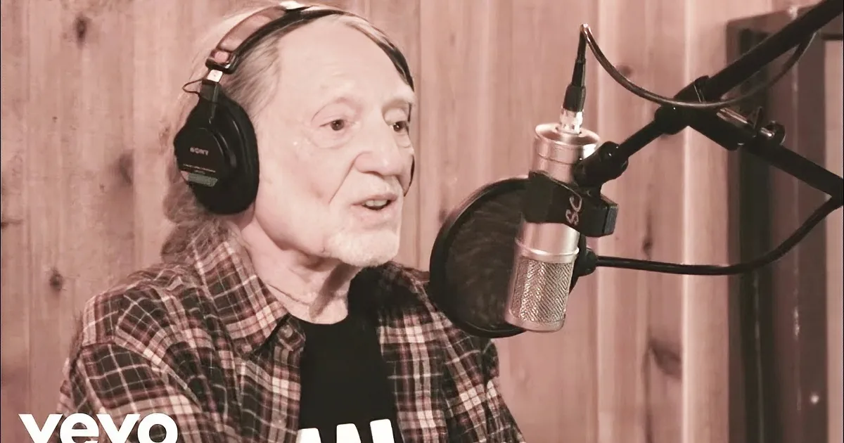 An older man wearing headphones and a plaid shirt is smiling in front of a microphone in a recording studio.