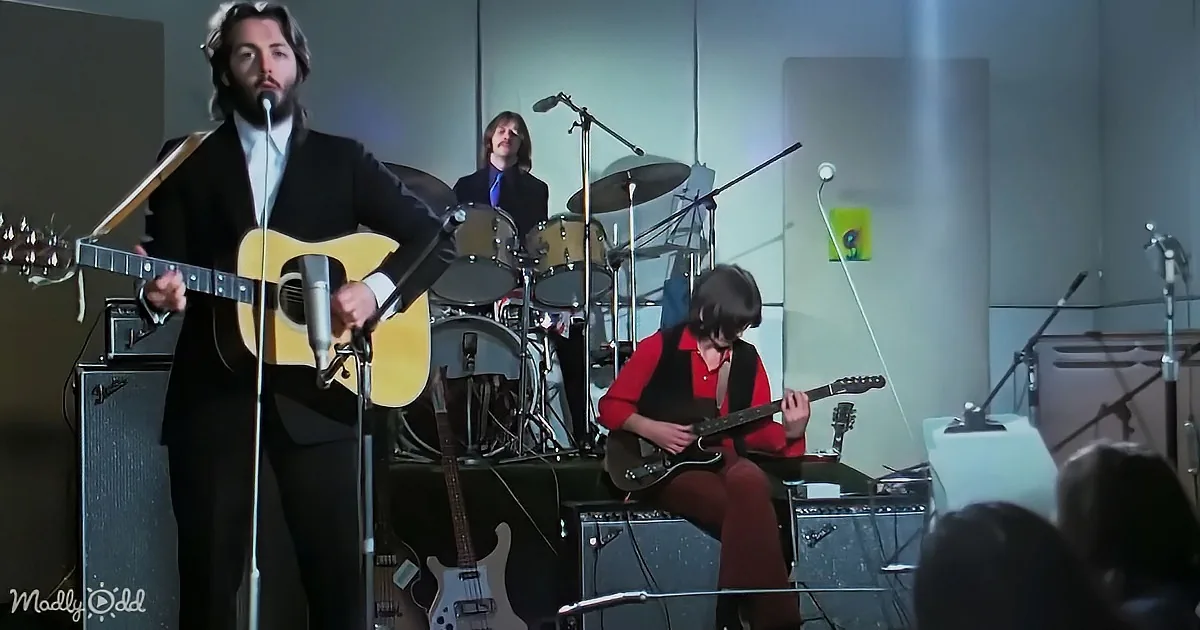 A man in a suit plays guitar on stage with a band, including a drummer and another guitarist.