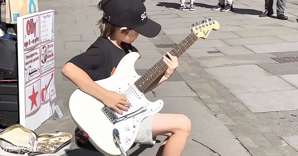 A young boy in a black cap plays a white electric guitar on a city sidewalk, with a sign and a tip jar nearby.
