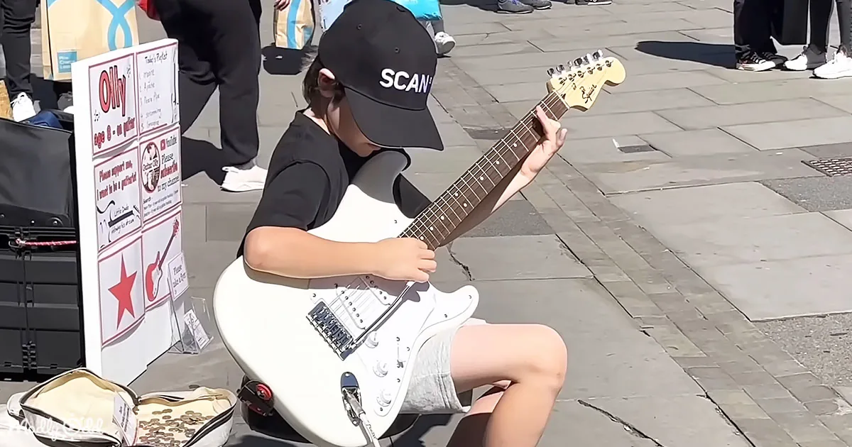 A young boy wearing a "SCAN" cap plays a white electric guitar on a city sidewalk.