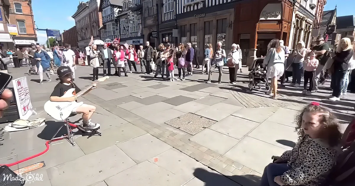 A young guitarist performs on a city street, surrounded by a crowd of spectators.