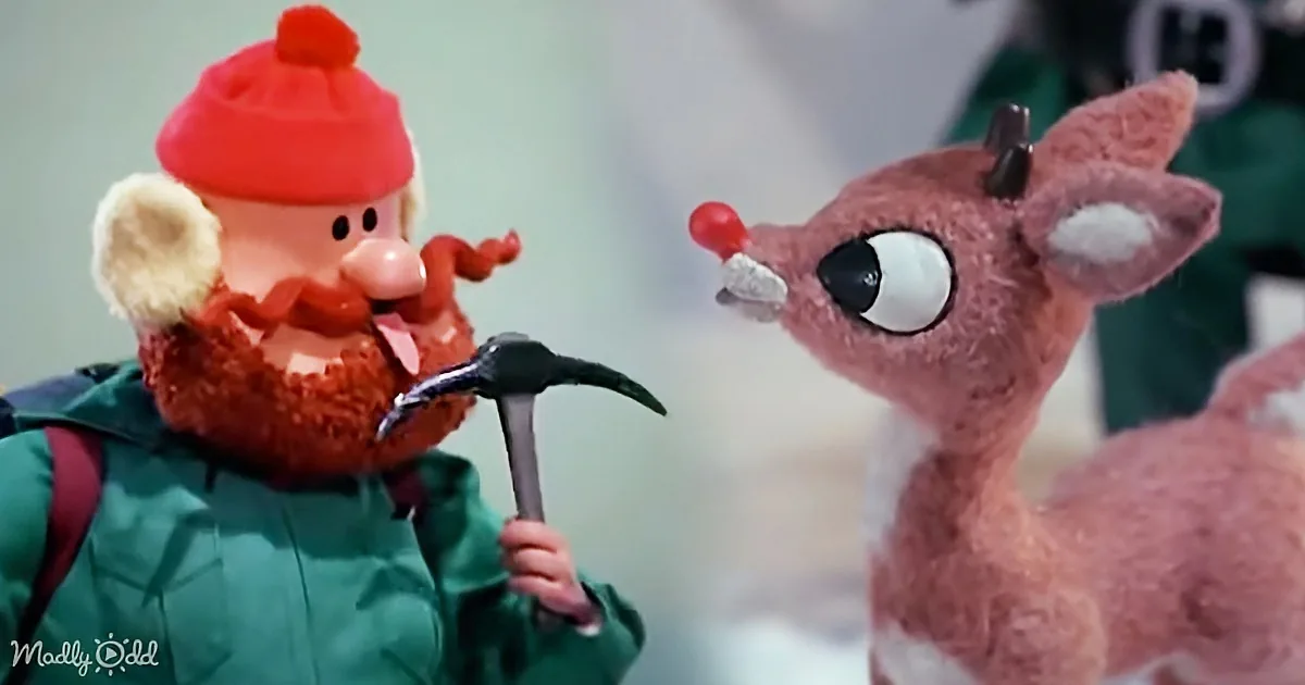 A bearded man in a red hat and green jacket holds a hammer, facing a surprised-looking reindeer with a red nose.