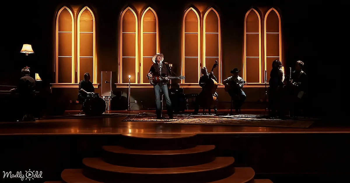 A man in a cowboy hat sings on stage with a band, illuminated by warm light from arched windows behind.
