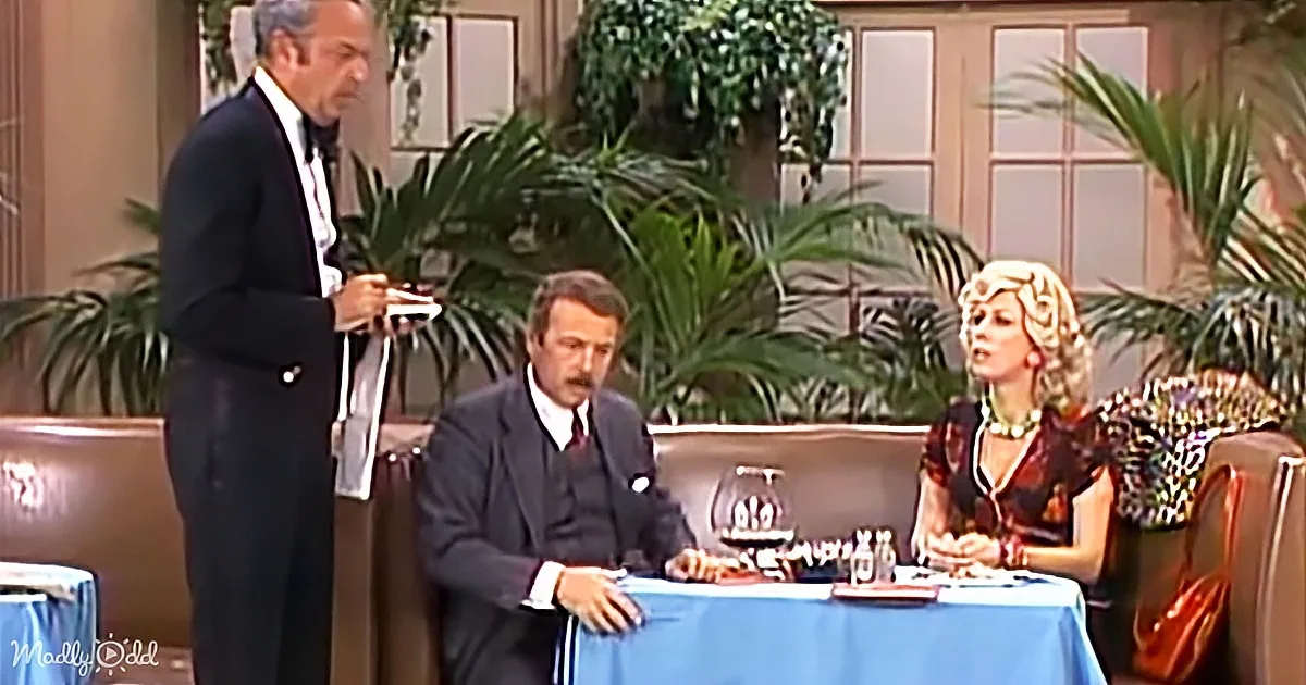 A man in a suit stands holding a microphone, addressing a seated couple at a table.