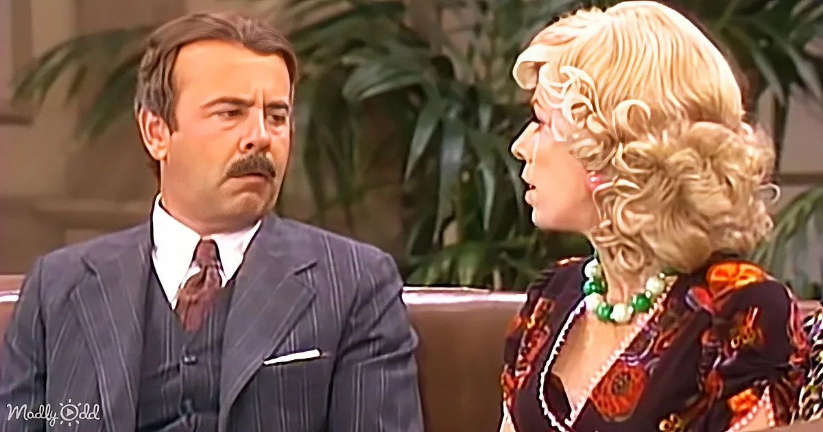 A man in a suit and a woman in a floral dress are seated on a couch, engaged in conversation.