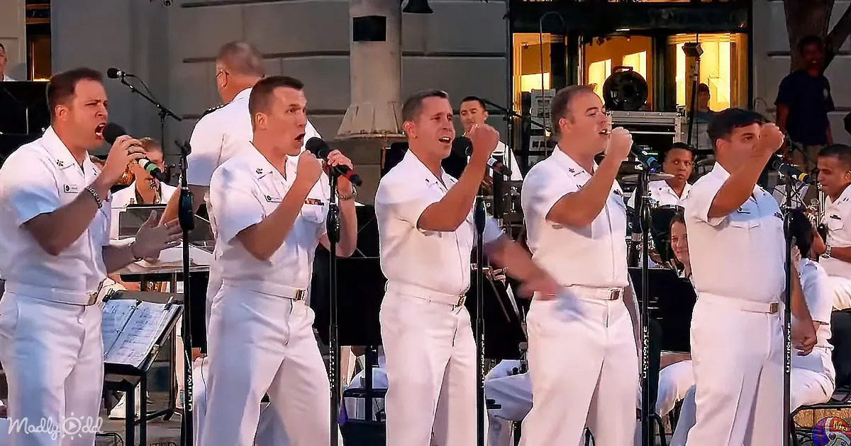 A group of men in white uniforms sing passionately on stage with microphones.