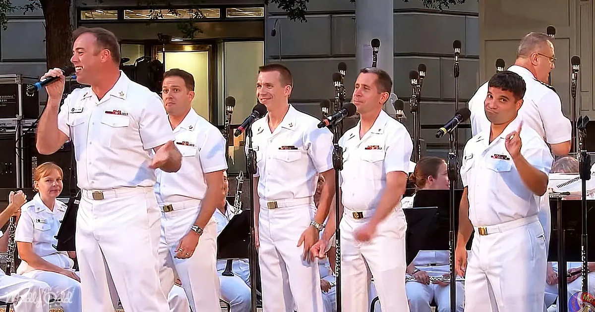 A group of men in white uniforms sing on stage with microphones, smiling and gesturing.