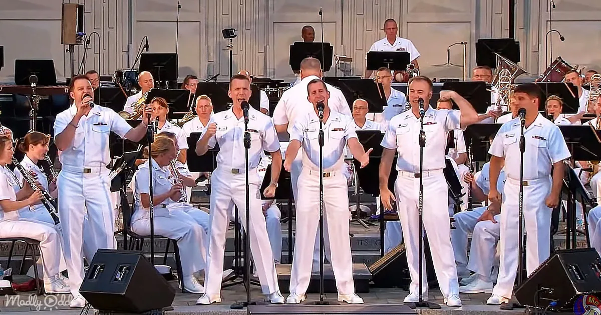 A group of uniformed men sing on stage with a band in the background.