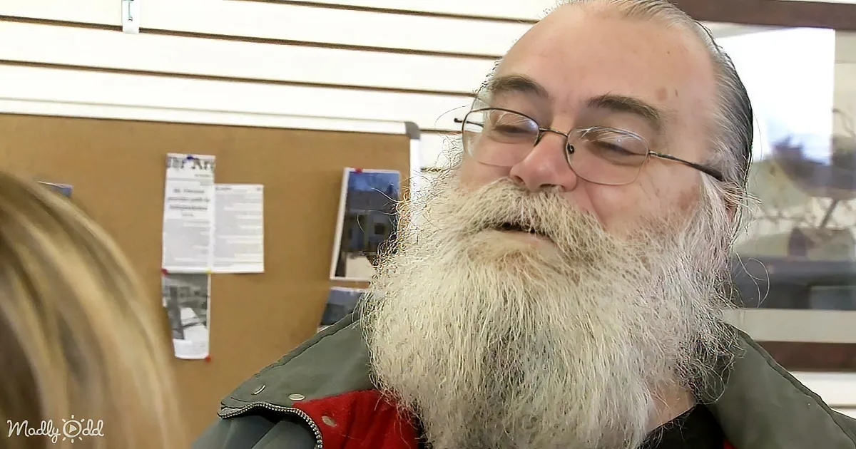 An older man with a long white beard and glasses smiles in front of a corkboard with papers and photos.
