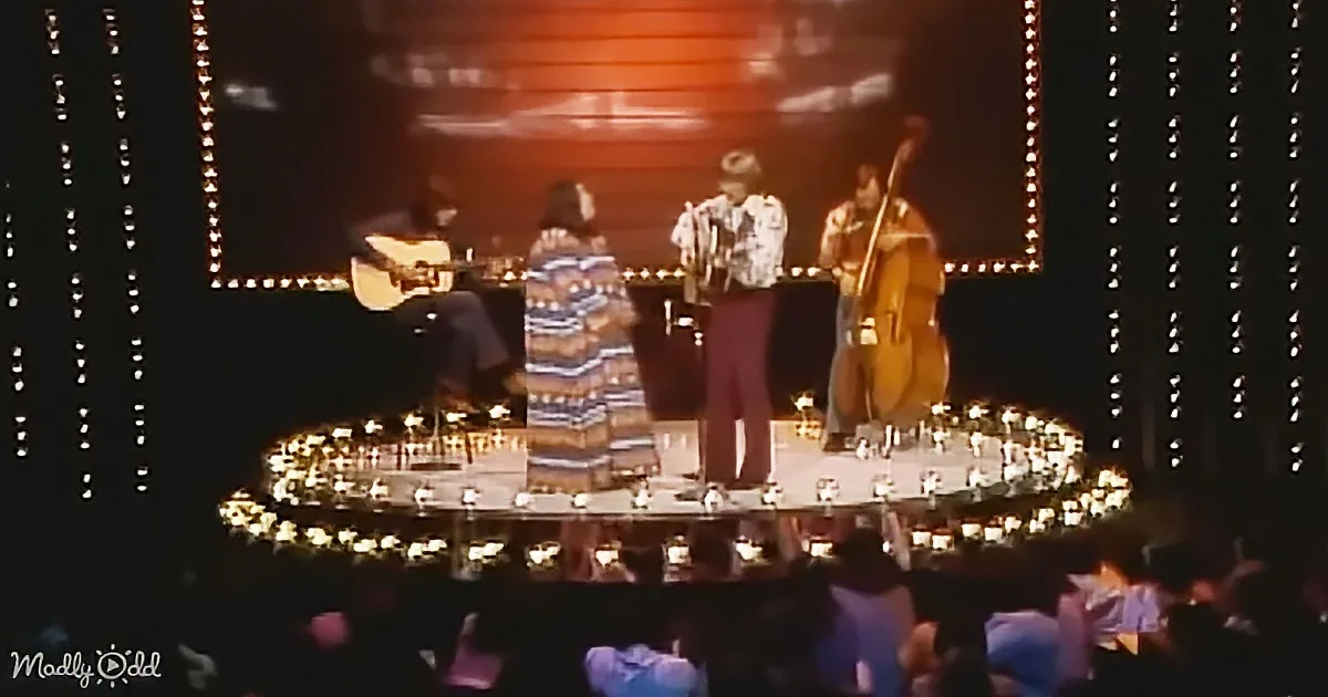 A band performs on stage with a guitarist, bassist, and drummer, surrounded by lights.